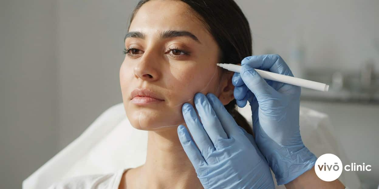 Gloved hands of a practitioner marking white assessment lines on a Middle Eastern woman’s jawline during a careful pre-treatment skin evaluation, no device in frame