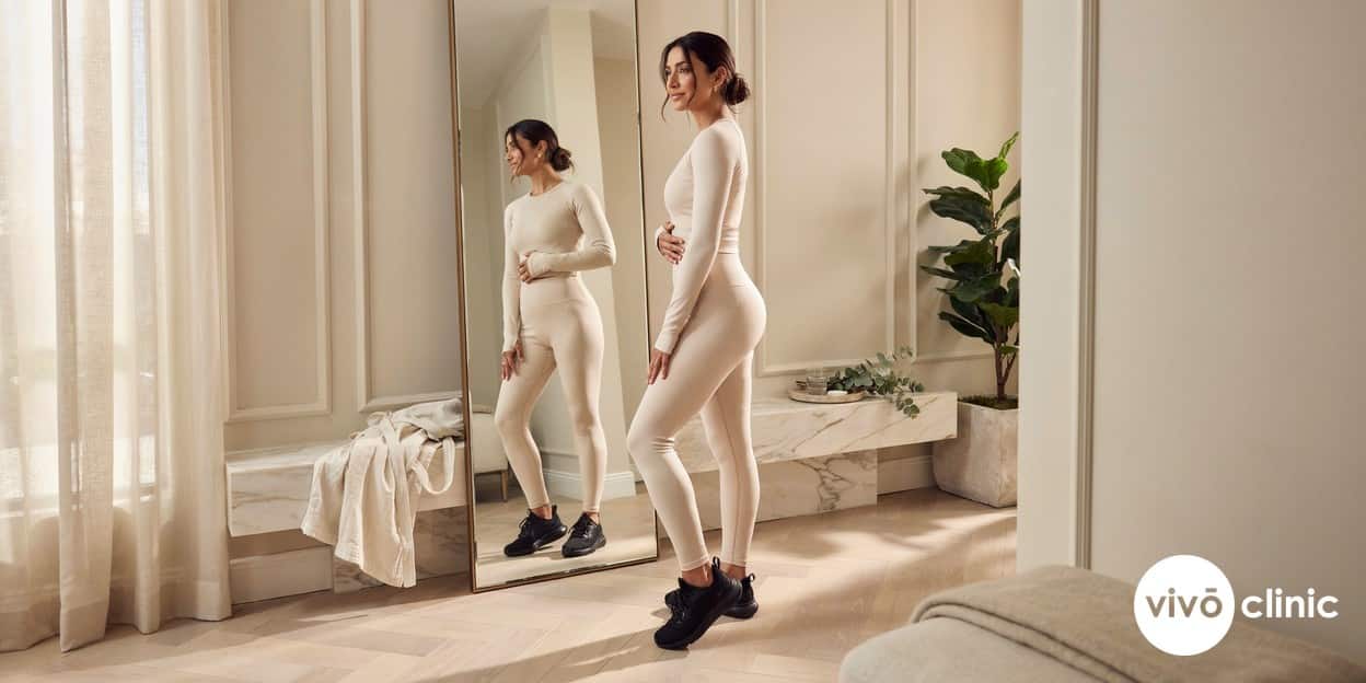 Middle Eastern woman in activewear assessing the smoother appearance of her thighs in a bright mirror after a course of cellulite treatment at VIVO Clinic Dubai