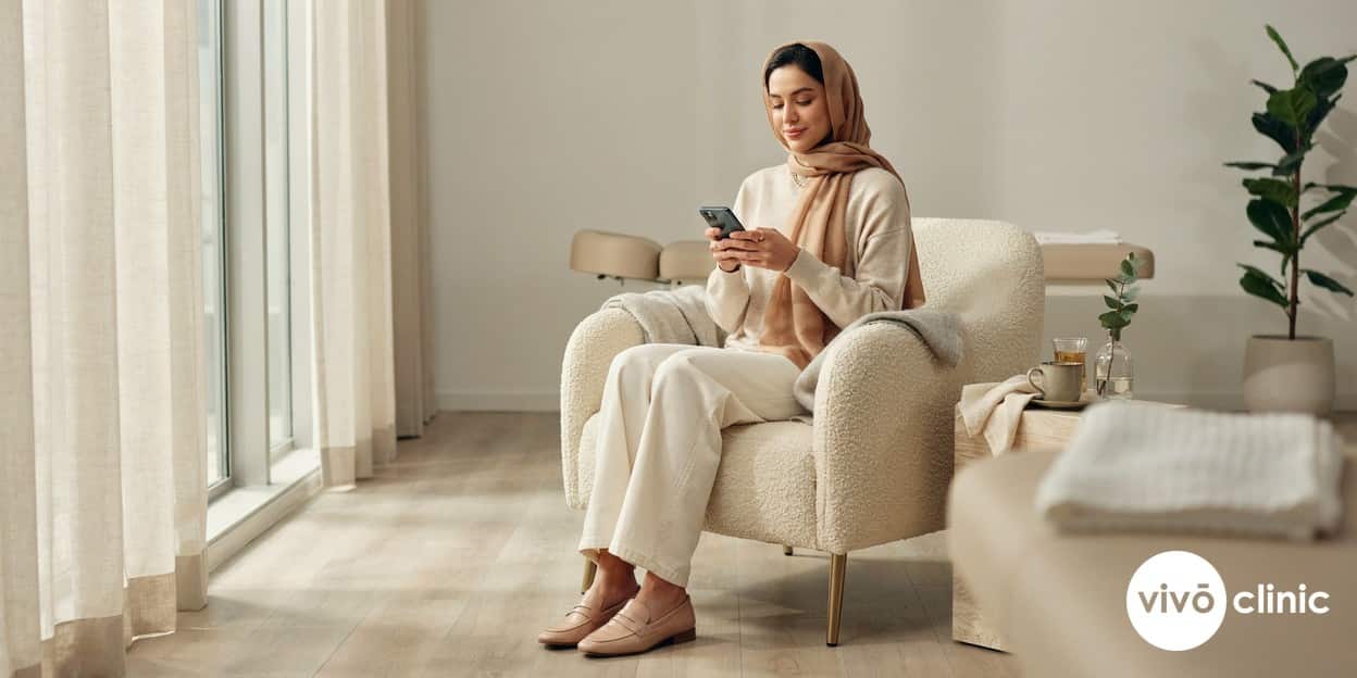 Middle Eastern woman in modest dress sitting calmly and reading on her phone during the comfortable middle phase of a fat freezing session in a premium Dubai clinic, no device, machine or handpiece anywhere in frame