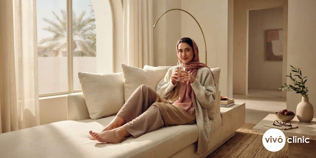 Middle Eastern woman smiling and resting at home with a glass of water after her fat freezing appointment at VIVO Clinic Dubai, relaxed and comfortable in modest clothing, no device, machine or handpiece anywhere in frame