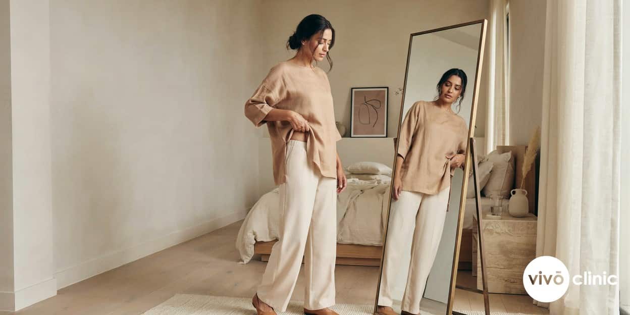 Middle Eastern woman in modest clothing pinching a small pocket of stubborn lower-abdomen fat in front of a mirror, illustrating the localised areas fat freezing is designed to contour