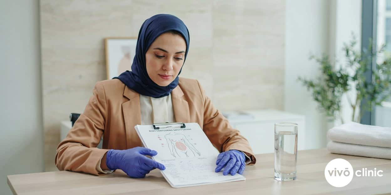 Practitioner in modest attire reviewing a body-contouring safety chart and aftercare notes on a clipboard in a calm consultation setting, no device or handpiece in frame
