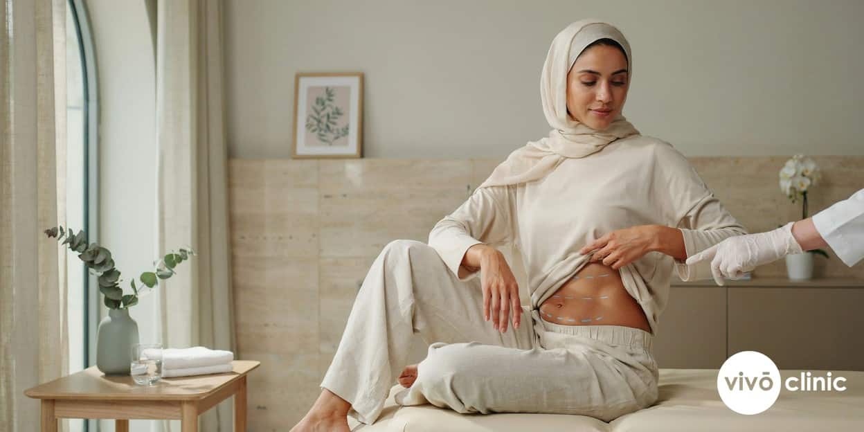 Middle Eastern woman in modest clothing with white cosmetic marking lines drawn on her abdomen during a body contouring consultation at VIVO Clinic Dubai, comparing fat freezing and ultrasound cavitation
