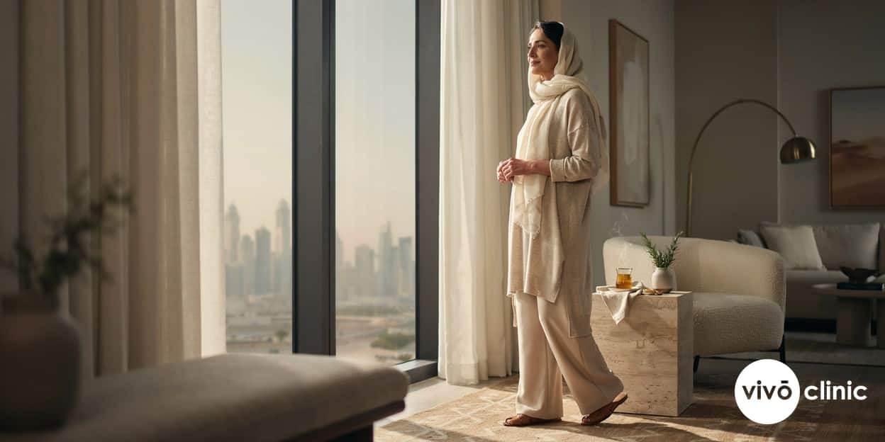 Middle Eastern woman in her fifties in modest, elegant home clothing looking calm and relaxed by a sunlit window, conveying renewed comfort and wellbeing during menopause