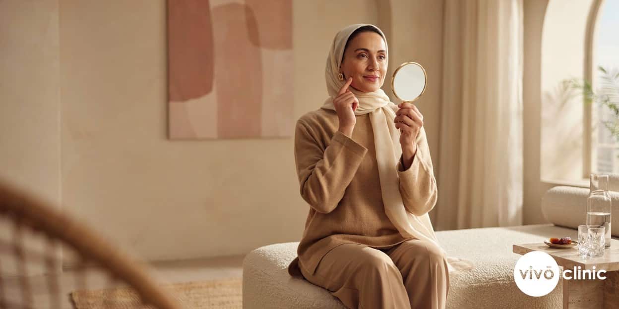 Middle Eastern woman in modest attire examining her firmer jawline in a handheld mirror at VIVO Clinic Dubai, three months after a radiofrequency facelift