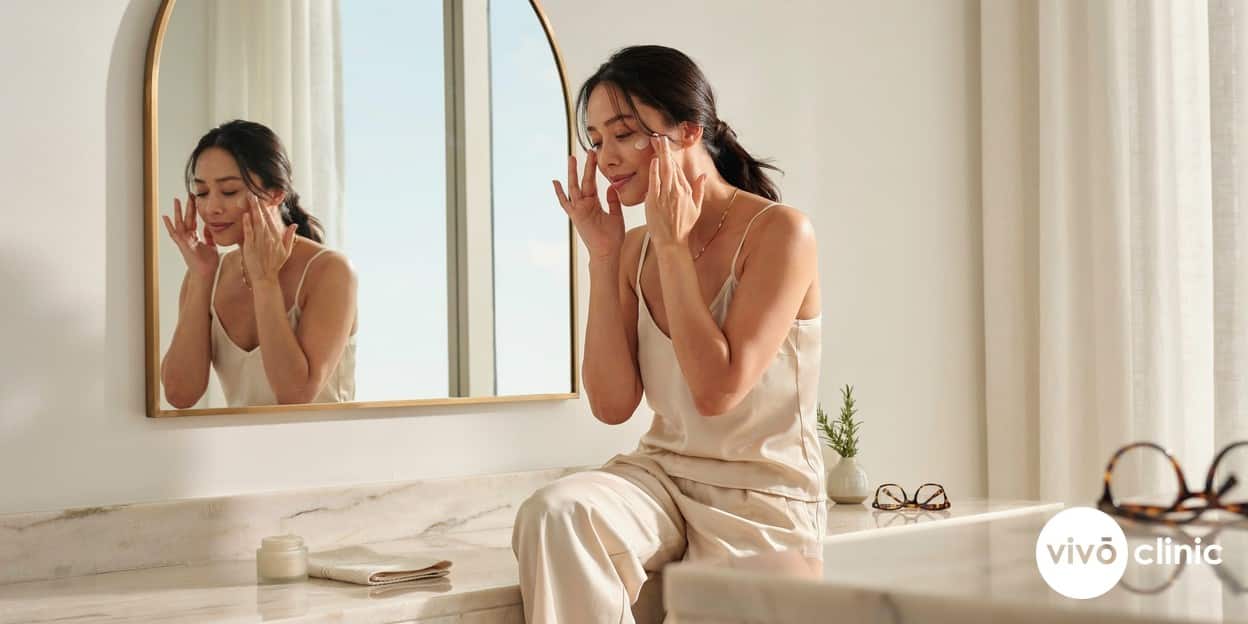 Middle Eastern woman applying sunscreen at a bright bathroom mirror as part of her HIFU facelift aftercare routine at VIVO Clinic Dubai, no device, machine or handpiece anywhere in frame