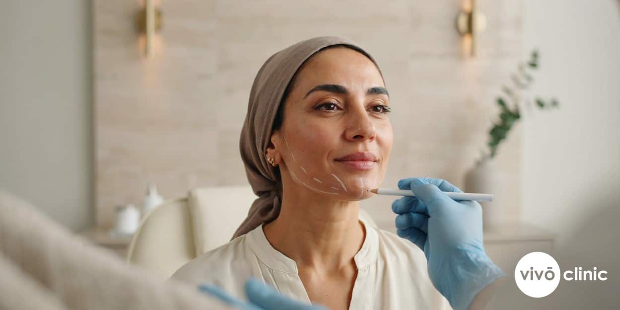 Middle Eastern woman in modest dress with white cosmetic marking lines drawn beneath her chin during an Aqualyx fat-dissolving consultation at VIVO Clinic Dubai