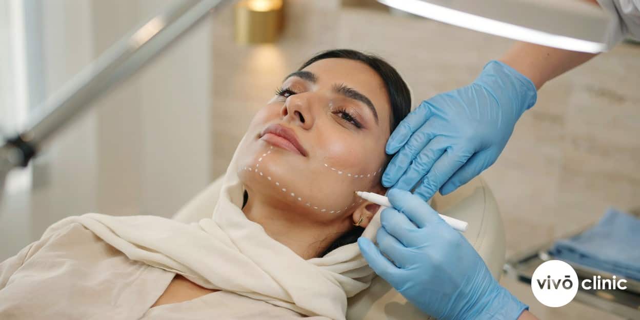Gloved hands of a practitioner drawing a white cosmetic injection-grid pattern onto the under-chin area of a Middle Eastern woman in modest attire at VIVO Clinic Dubai, demonstrating careful intralipotherapy mapping for safe fat dissolving, no device, machine or handpiece anywhere in frame