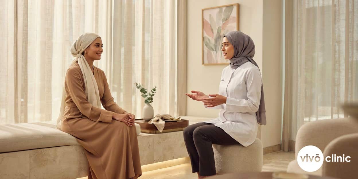 Middle Eastern woman in modest dress smiling during a relaxed post-consultation conversation with a female practitioner in a premium, calm VIVO Clinic Dubai treatment room, no device, machine or handpiece anywhere in frame