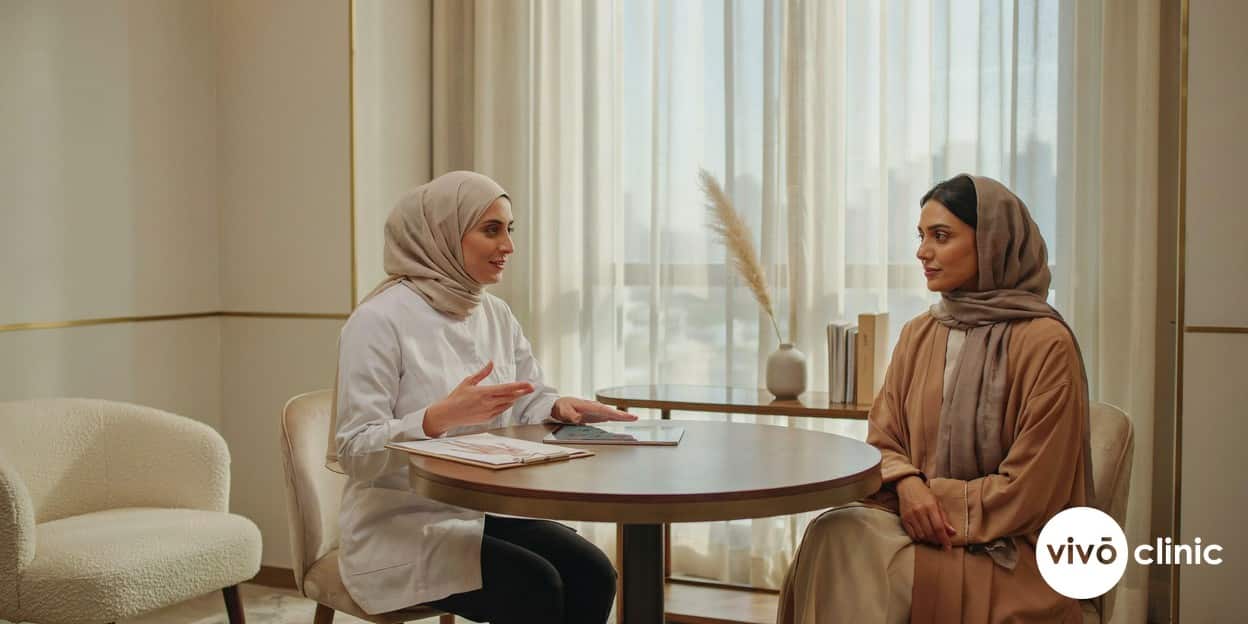 Middle Eastern woman in modest dress during an Aqualyx fat dissolving consultation with a gloved practitioner at VIVO Clinic Dubai, reviewing safety and suitability before treatment