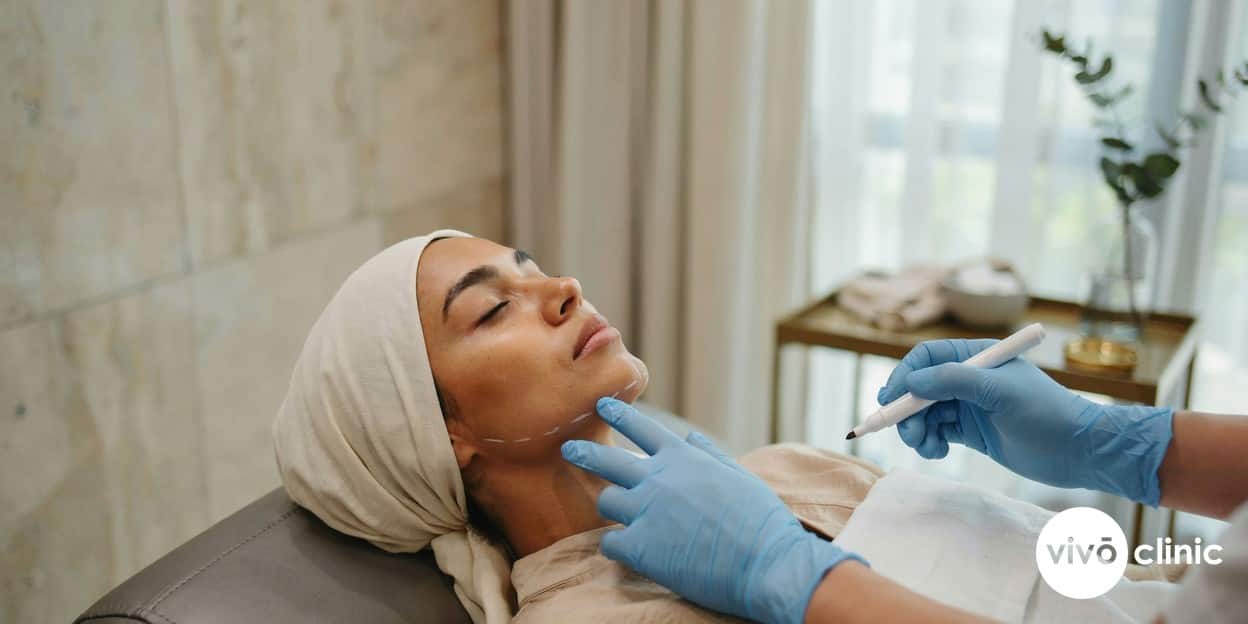 Practitioner in modest attire gently marking white cosmetic guide lines along the jawline of a relaxed Middle Eastern woman during a HIFU facelift consultation at VIVO Clinic Dubai, no device, machine or handpiece anywhere in frame