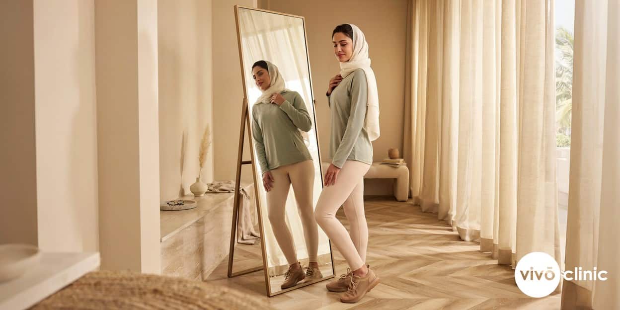 Middle Eastern woman in modest activewear looking confidently at her reflection in a bright Dubai clinic after a course of non-surgical body contouring, no device, machine or handpiece anywhere in frame