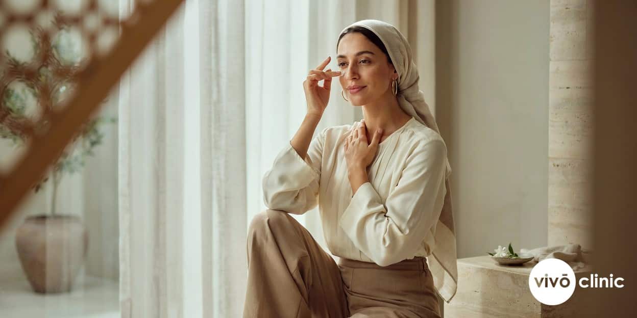 Middle Eastern woman applying high-SPF sunscreen around her healed, brighter eye area in front of a bright window, illustrating sun protection after a plasma eye lift