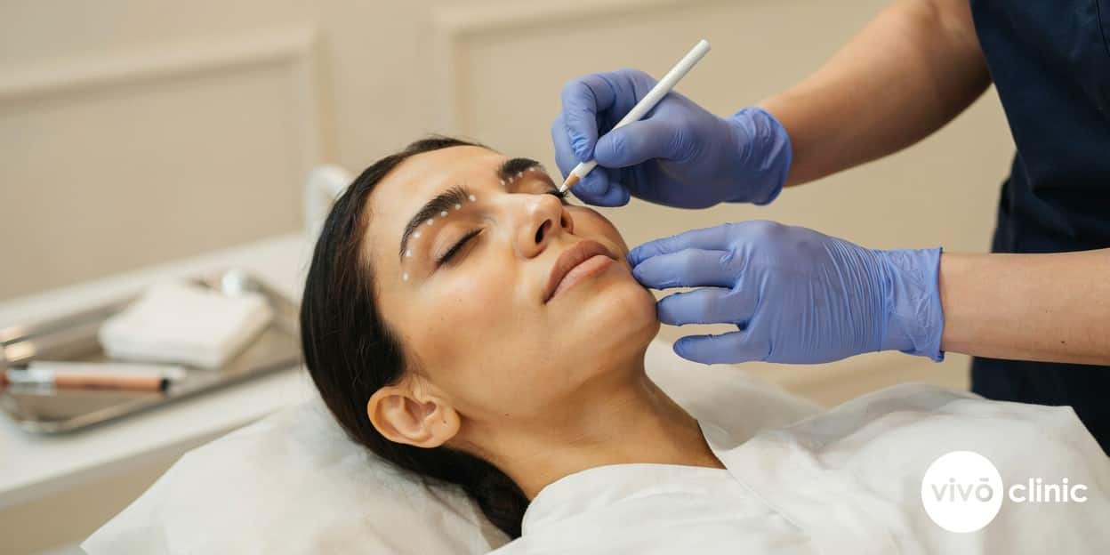 Practitioner in modest attire wearing gloves, gently marking fine white assessment dots along the upper eyelid crease of a Middle Eastern woman during a plasma eye lift consultation, no device, machine or handpiece anywhere in frame