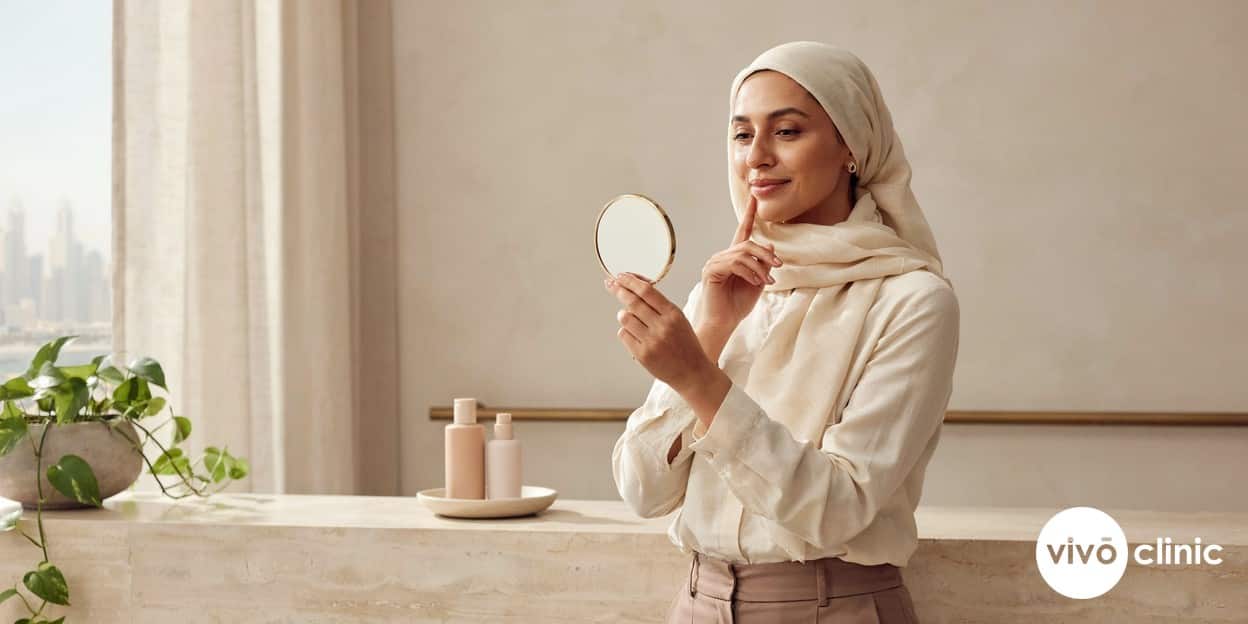 Middle Eastern woman examining her firmer, smoother jawline and cheek in a mirror after a course of RF skin tightening at VIVO Clinic Dubai