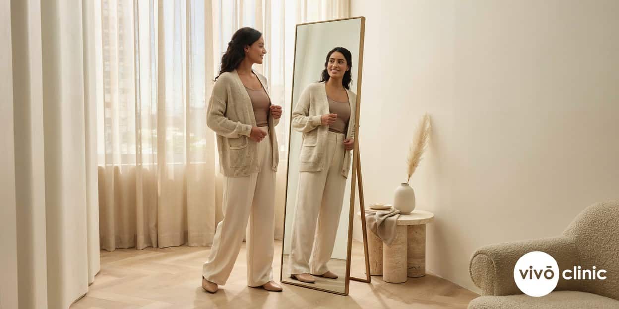 Middle Eastern woman in modest clothing smiling as she checks her flatter abdomen profile in a mirror at a premium VIVO Clinic Dubai consultation room