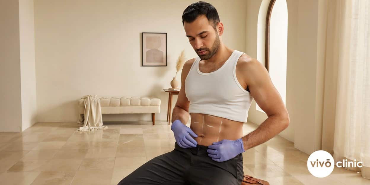 Middle Eastern man in modest clothing having his abdomen assessed with white cosmetic marking lines by a gloved practitioner during a fat freezing suitability consultation, illustrating that men and abdominal treatment carry a higher PAH risk