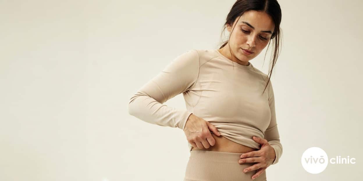 Middle Eastern woman in modest activewear pinching a localised lower-abdominal fat roll, illustrating the pinchable subcutaneous fat that makes an ideal fat freezing candidate