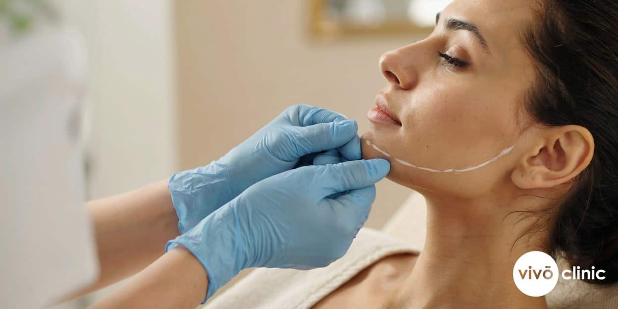 Close-up of a practitioner’s gloved hands performing a gentle pinch test on a Middle Eastern woman’s jawline to assess skin elasticity before Aqualyx, white cosmetic marking line visible, no device or handpiece in frame
