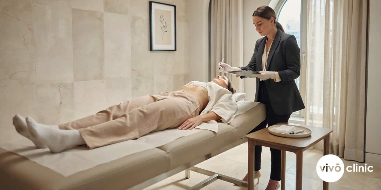 Practitioner in modest attire wearing gloves, drawing white cosmetic marking lines on a Middle Eastern woman’s abdomen and pausing to note a contraindication, no device, machine or handpiece anywhere in frame